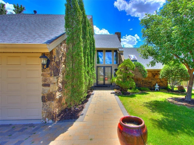 view of exterior entry featuring an attached garage, a shingled roof, a yard, stone siding, and french doors