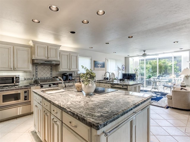 kitchen featuring light tile patterned flooring, ceiling fan, dark stone counters, and an island with sink