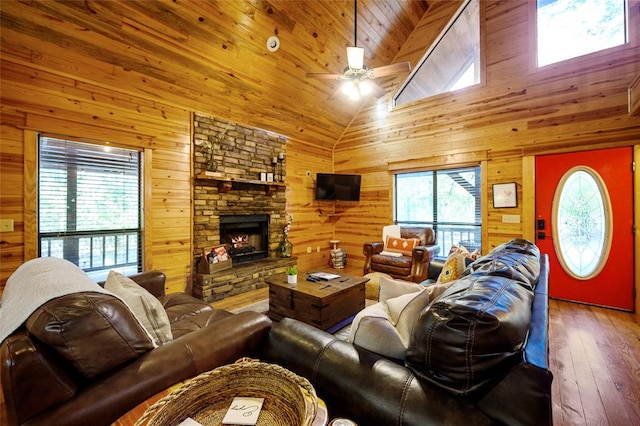 living room featuring ceiling fan, wooden walls, high vaulted ceiling, hardwood / wood-style floors, and a stone fireplace