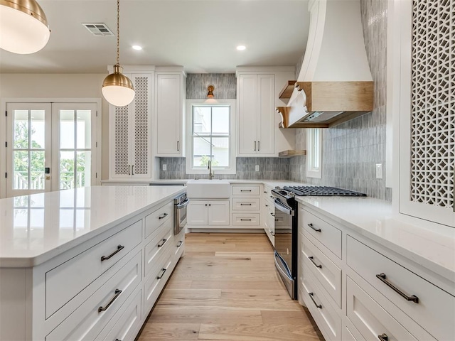 kitchen with custom exhaust hood, light hardwood / wood-style flooring, stainless steel gas stove, decorative light fixtures, and white cabinetry