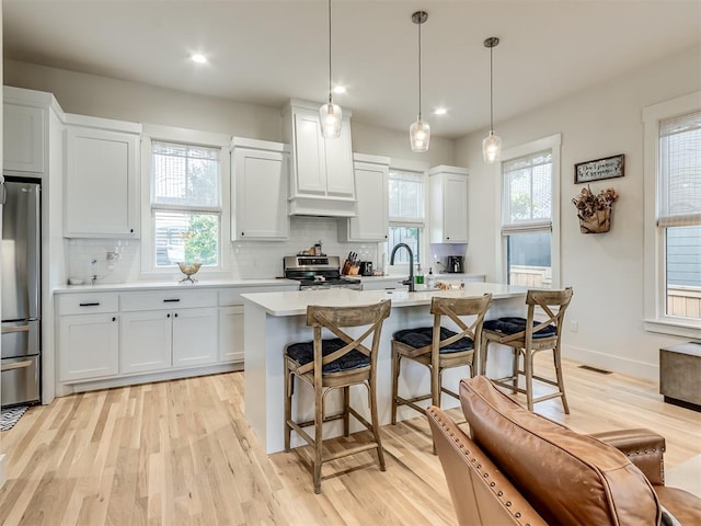 kitchen with appliances with stainless steel finishes, light wood-type flooring, a kitchen island with sink, pendant lighting, and white cabinets