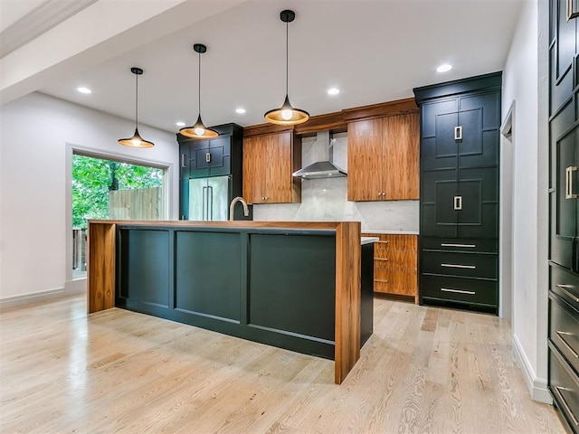 kitchen with light wood-type flooring, a kitchen island with sink, tasteful backsplash, wall chimney exhaust hood, and pendant lighting