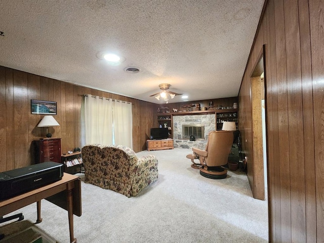 carpeted living room featuring a stone fireplace, wooden walls, ceiling fan, and a textured ceiling