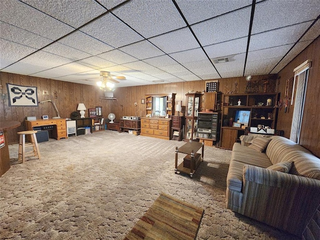 living room featuring ceiling fan, a drop ceiling, carpet floors, and wooden walls