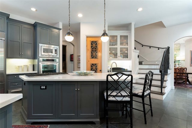 kitchen featuring gray cabinets, an island with sink, appliances with stainless steel finishes, tasteful backsplash, and decorative light fixtures