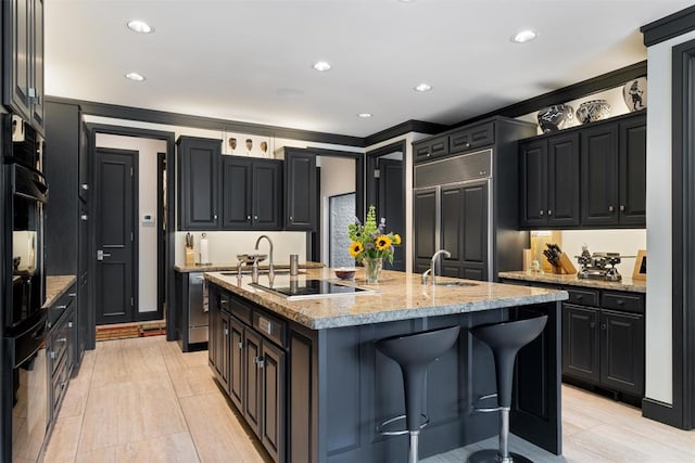 kitchen featuring light stone countertops, sink, paneled refrigerator, an island with sink, and black electric stovetop