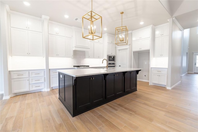 kitchen with white cabinetry, hanging light fixtures, an island with sink, built in microwave, and light wood-type flooring