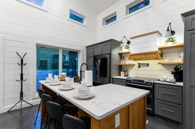 kitchen with light stone countertops, stainless steel refrigerator with ice dispenser, electric range, gray cabinets, and an island with sink