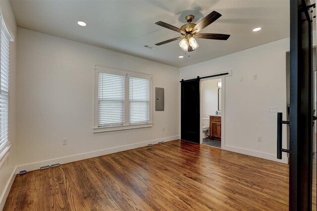 unfurnished bedroom with ceiling fan, a barn door, hardwood / wood-style flooring, connected bathroom, and electric panel