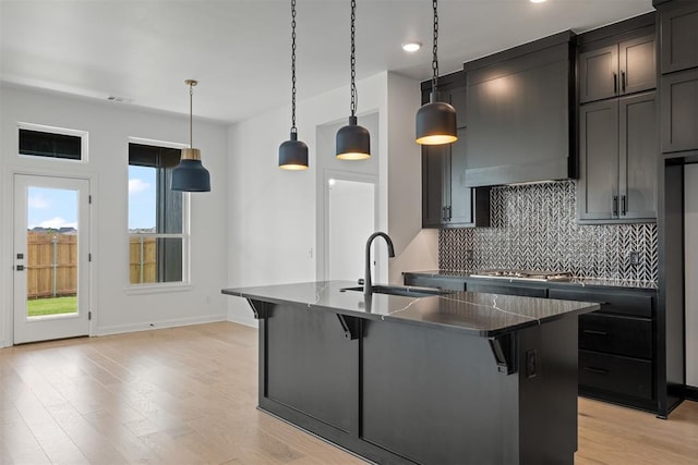 kitchen with sink, wall chimney range hood, pendant lighting, light hardwood / wood-style flooring, and an island with sink