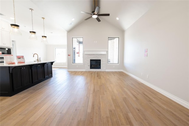 unfurnished living room with ceiling fan, sink, a stone fireplace, high vaulted ceiling, and light hardwood / wood-style floors