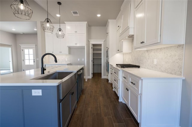 kitchen with white cabinets, appliances with stainless steel finishes, custom exhaust hood, hanging light fixtures, and a center island with sink