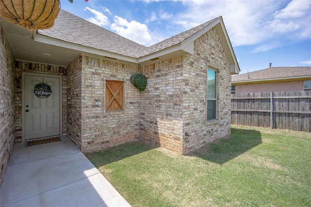 view of exterior entry with a yard, brick siding, a shingled roof, and fence