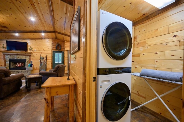 clothes washing area with stacked washer / drying machine, a stone fireplace, wooden ceiling, and wooden walls