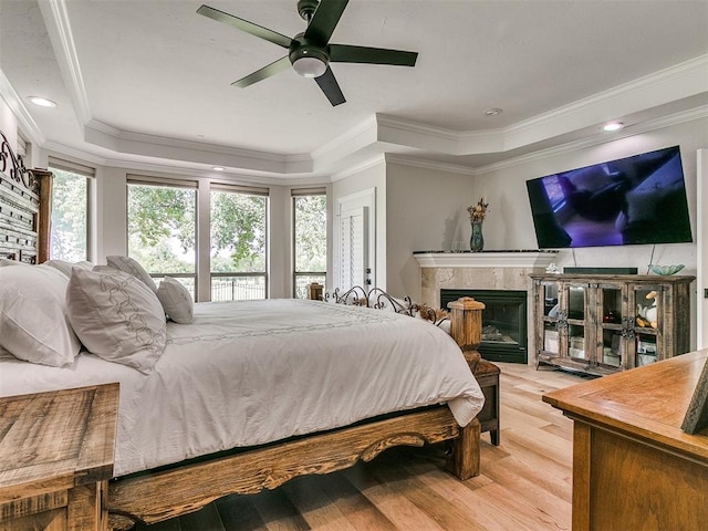 bedroom featuring ceiling fan, a premium fireplace, light hardwood / wood-style flooring, crown molding, and a tray ceiling