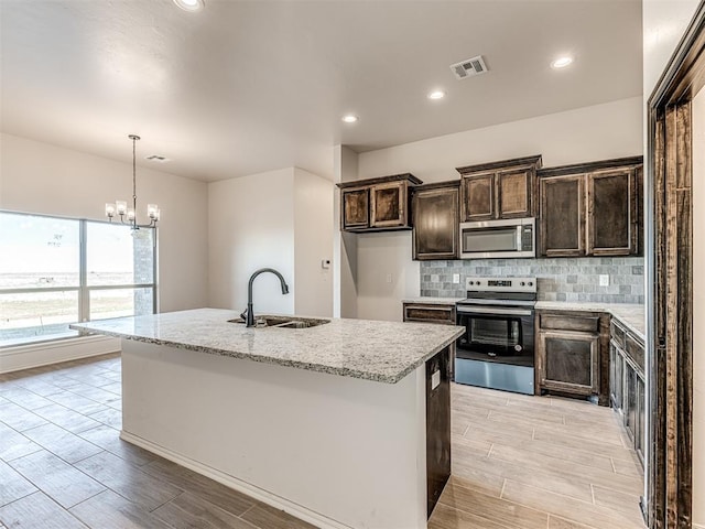 kitchen with appliances with stainless steel finishes, light stone counters, a kitchen island with sink, sink, and a notable chandelier