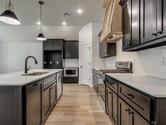 kitchen with appliances with stainless steel finishes, light wood-type flooring, custom range hood, pendant lighting, and a center island with sink