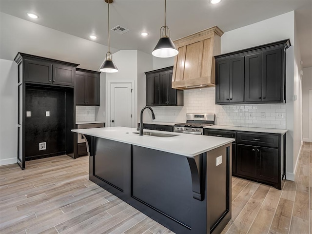 kitchen with light wood-type flooring, premium range hood, gas stove, a kitchen island with sink, and sink