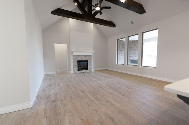 unfurnished living room with a stone fireplace, high vaulted ceiling, ceiling fan, beam ceiling, and light hardwood / wood-style floors