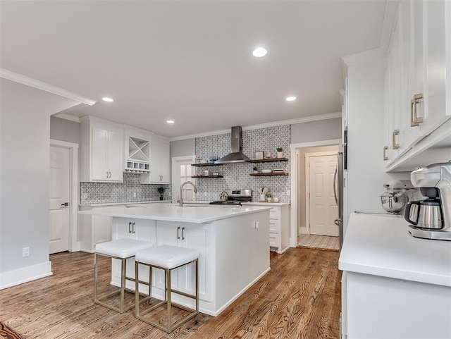 kitchen with ornamental molding, wall chimney range hood, a center island with sink, white cabinets, and hardwood / wood-style floors