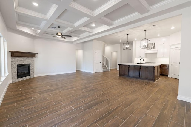 unfurnished living room featuring coffered ceiling, a stone fireplace, sink, beam ceiling, and ceiling fan with notable chandelier