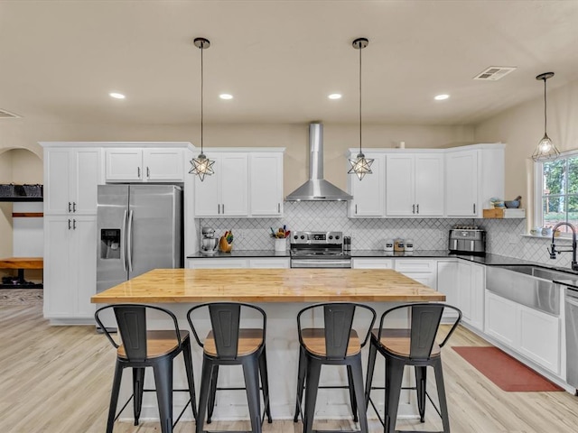 kitchen featuring white cabinetry, wall chimney range hood, light hardwood / wood-style flooring, decorative light fixtures, and appliances with stainless steel finishes