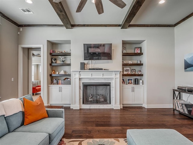 living room with dark wood-type flooring, built in features, a premium fireplace, ceiling fan, and beamed ceiling