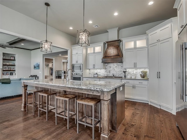 kitchen with white cabinetry, appliances with stainless steel finishes, custom range hood, and a center island with sink