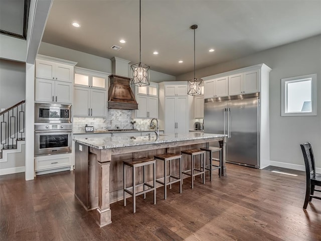 kitchen featuring premium range hood, decorative light fixtures, an island with sink, white cabinets, and built in appliances