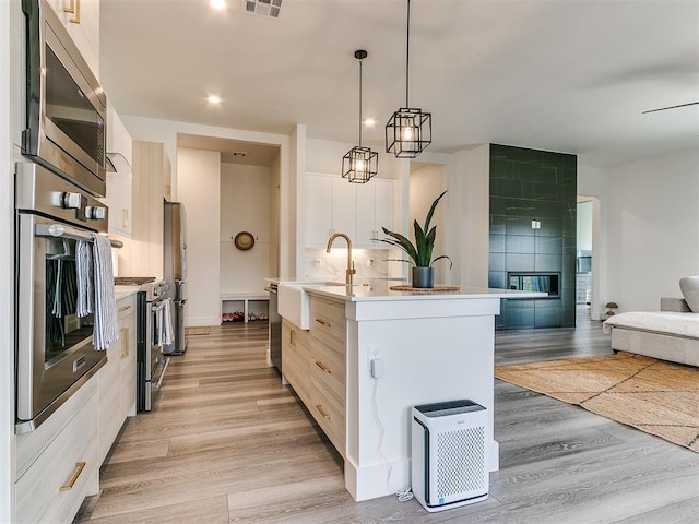 kitchen featuring stainless steel appliances, backsplash, light hardwood / wood-style floors, decorative light fixtures, and a center island with sink