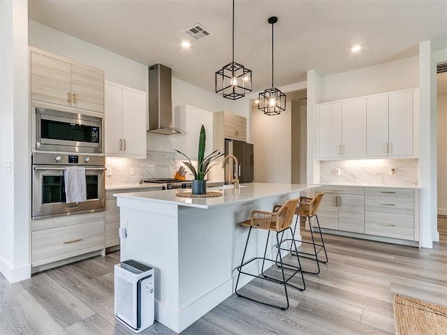 kitchen with white cabinetry, a center island with sink, wall chimney range hood, and appliances with stainless steel finishes