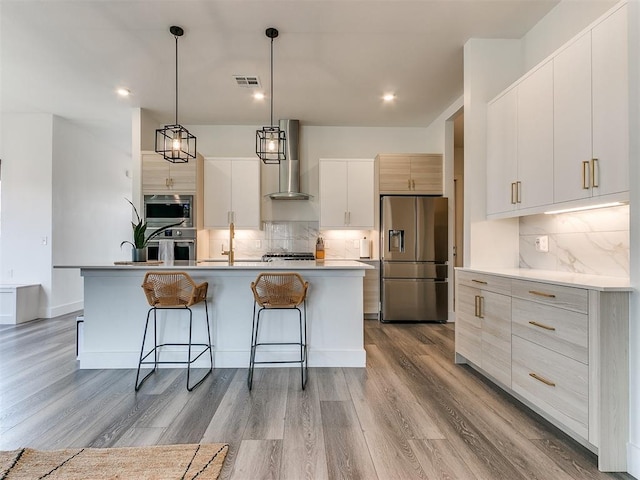 kitchen featuring appliances with stainless steel finishes, wall chimney exhaust hood, a kitchen island with sink, pendant lighting, and white cabinets