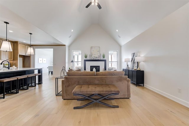 living room featuring high vaulted ceiling, sink, a brick fireplace, ceiling fan, and light wood-type flooring