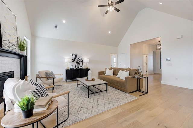 living room featuring a brick fireplace, high vaulted ceiling, ceiling fan with notable chandelier, and light wood-type flooring