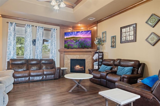 living room featuring ceiling fan, a raised ceiling, dark hardwood / wood-style floors, a tiled fireplace, and ornamental molding