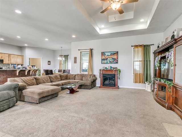 living room featuring carpet flooring, ceiling fan with notable chandelier, a tray ceiling, and a brick fireplace