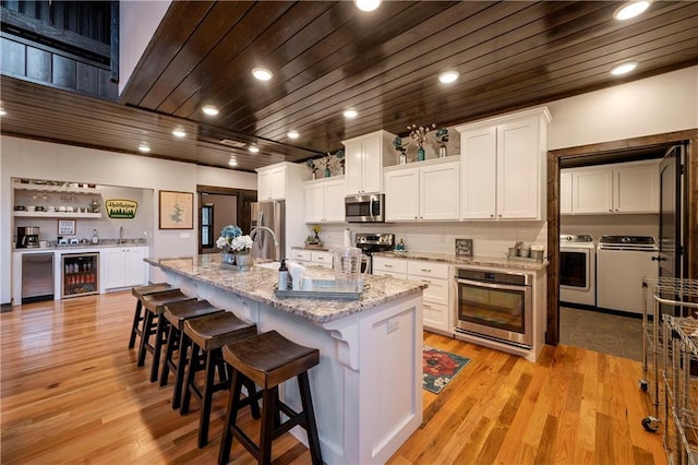 kitchen with washing machine and dryer, an island with sink, white cabinetry, stainless steel appliances, and beverage cooler