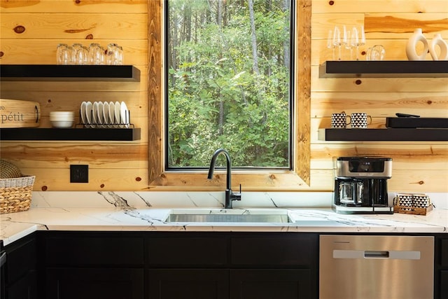 kitchen featuring stainless steel dishwasher, wood walls, light stone countertops, and sink