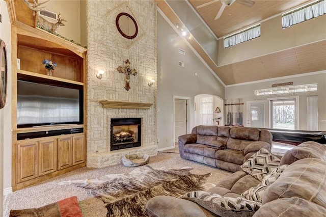 living room featuring ceiling fan, a stone fireplace, high vaulted ceiling, crown molding, and light colored carpet