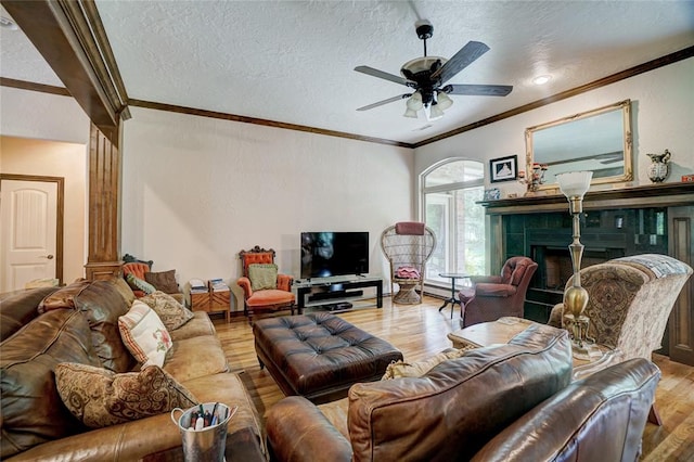 living room featuring light wood-type flooring, a textured ceiling, ceiling fan, crown molding, and a tiled fireplace