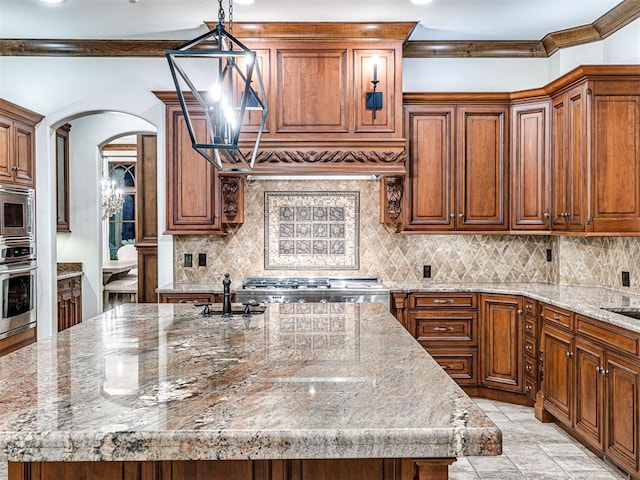 kitchen with appliances with stainless steel finishes, backsplash, light stone counters, a center island with sink, and hanging light fixtures