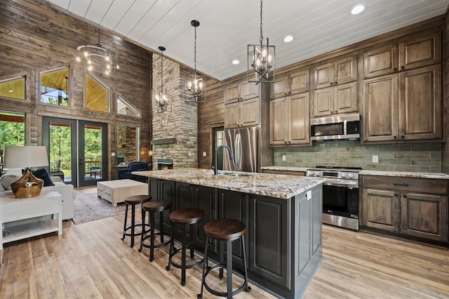 kitchen with appliances with stainless steel finishes, a kitchen island with sink, wooden walls, high vaulted ceiling, and light hardwood / wood-style floors