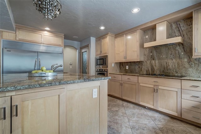 kitchen featuring built in appliances, light brown cabinets, light stone countertops, and wall chimney range hood