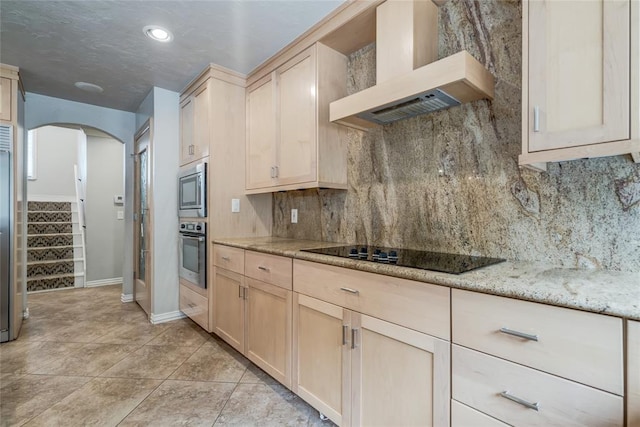 kitchen with appliances with stainless steel finishes, backsplash, light stone counters, wall chimney range hood, and light brown cabinets