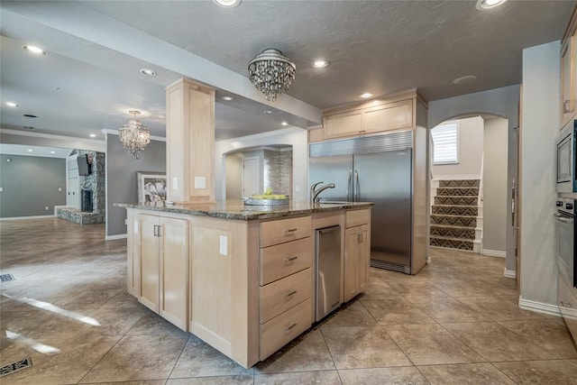 kitchen with a stone fireplace, light brown cabinetry, an island with sink, and built in appliances