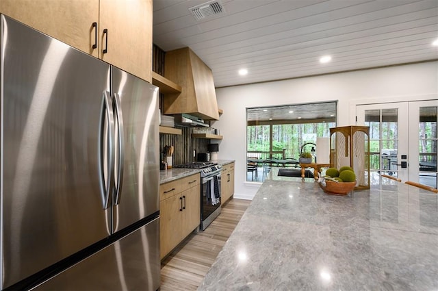 kitchen featuring light brown cabinets, light stone counters, premium range hood, light hardwood / wood-style floors, and appliances with stainless steel finishes