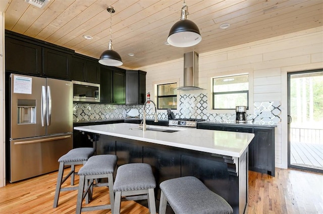 kitchen featuring a kitchen island with sink, wall chimney exhaust hood, hanging light fixtures, and appliances with stainless steel finishes