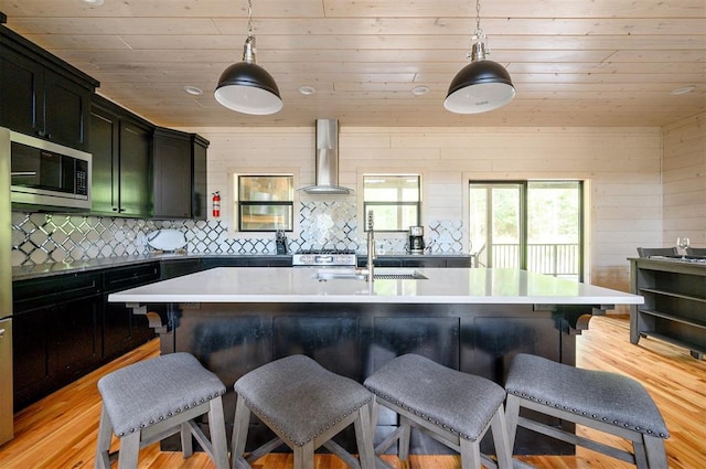 kitchen with stainless steel microwave, an island with sink, pendant lighting, and wall chimney range hood