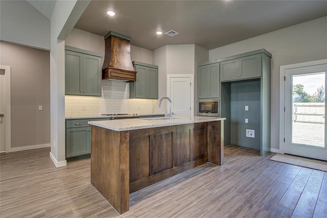 kitchen featuring premium range hood, light stone counters, a kitchen island with sink, built in microwave, and sink