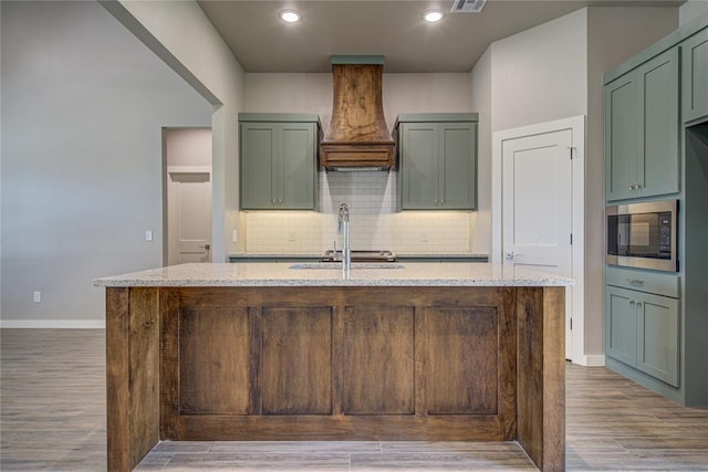 kitchen featuring built in microwave, light stone countertops, decorative backsplash, a kitchen island with sink, and custom exhaust hood